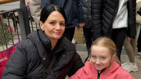 Gillian has black hair tied back and is wearing a black coat. She is sitting beside her daughter who has ginger hair and wearing a pink coat.
