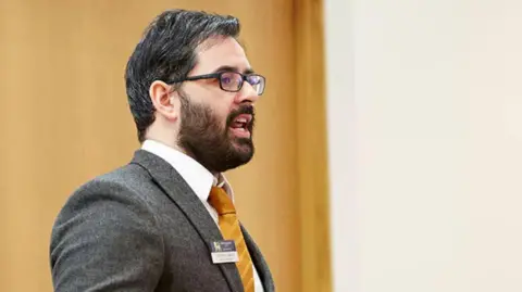 A man in a grey jacket, white shirt and yellow tie with dark hair, a beard and dark framed glasses