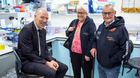 Dr Johnathan Cooper-Knock is pictured dressed in a navy suit seated on a chair in a university laboratory.  Standing to his right are Geoff and Irene Burrows. All three are smiling at the camera.