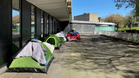 A collection of green and red tents pitched next to a grey concrete building