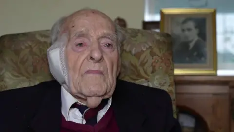 A front view of Roy Daines sat in a chair, looking slightly off camera at a reporter. He has grey hair on the sides of his head and is wearing a white shirt with a navy and red tie, a burgundy jumper and a black jacket. He has a bandage over his right ear.