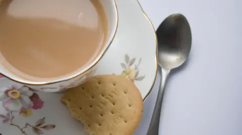 A cup of tea on a saucer, with a half eaten rich tea biscuit on the side. There is also a teaspoon next to the saucer.