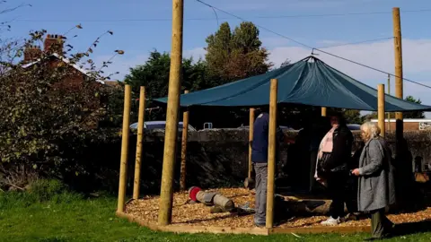 The Bay The new wildlife shelter in Preesall, Lancashire. Woodchip on the floor - a canopy in the centre of wooden posts - people gather around -  a house is in background