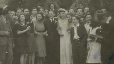 Black and white picture of people standing by the garden steps of the castle. Among the people is a bride and groom, wearing a wedding dress and suit