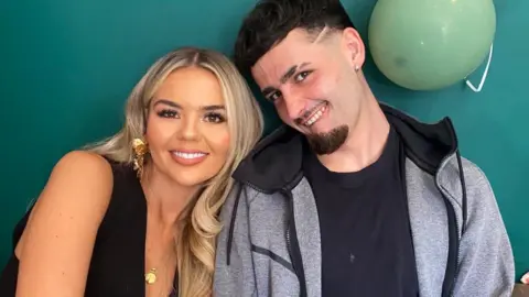 A woman with long blond hair and a black dress is sat with a man with short black hair, a chin beard and moustache dressed in a grey jacket and black t-shirt. They are sat against a green wall with a green balloon pinned to it.