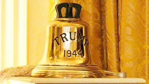 PA Media A gold bell from the HMS Trump placed on a white stand with gold curtains visible behind it.