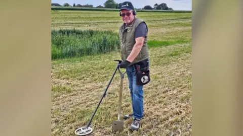 Steve Hickman Steve Hickman, a man standing in a green field with a shovel and a metal detector. He is looking directly at the camera and is wearing blue jeans, a blue T-shirt, a khaki gilet, a pair of sunglasses and a black baseball cap.