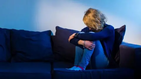 Blonde woman sits in a dark room, cradling her legs with her arms.