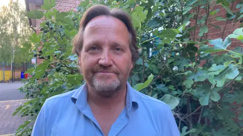 Matt Dormer is pictured outside next to a large bush. He has long brown swept back hair, a beard and is wearing a light blue shirt.