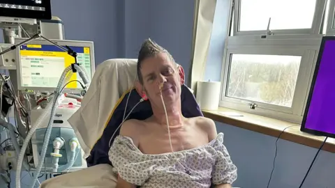 Kevin Heard A man sitting up in a hospital bed with tubes sticking out of his nose and ears with a medical machine behind him.