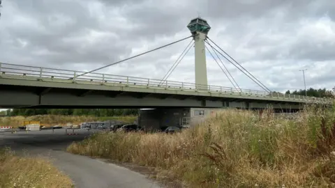 BBC/Richard Edwards This picture shows a bridge across the River Ouse, on the outskirts of the town of Selby. The bridge is painted light green. A control tower, which contains the mechanics needed to open and close the bridge, is in the middle of the image. Eight supporting cables connect the tower to the bridge itself.