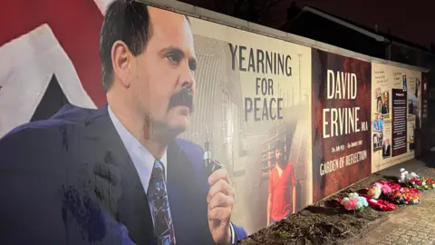 A memorial on a wall. It shows a side profile of David Ervine holding a pipe. One board says yearning for peace. One says David Ervine Garden of Reflection. Flowers are on the ground. 