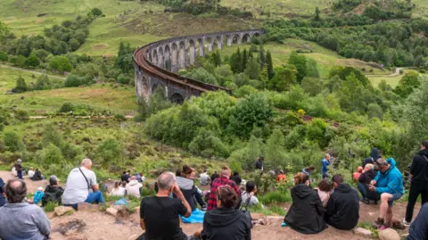 Getty Images The people are sitting on an area of bare ground on the hillside. The viaduct is a long bridge-like structure made up of a series of arches. The viaduct is set in a Highland landscape of hills, moorland and woods.