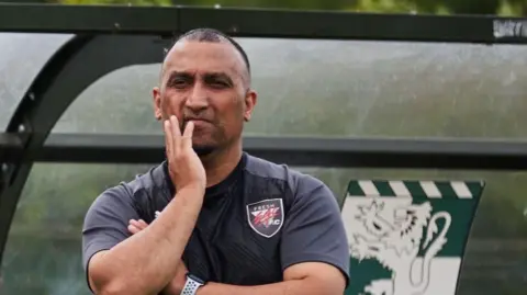 George Akhtar George Akhtar with very short dark hair with his right hand on his right cheek. He is wearing a blue T-shirt and a blue football club emblem. There is a lion logo behind him.