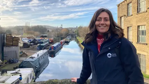 A woman with dark, long hair and wearing dark blue coat standing on a bridge with a canal running behind her 