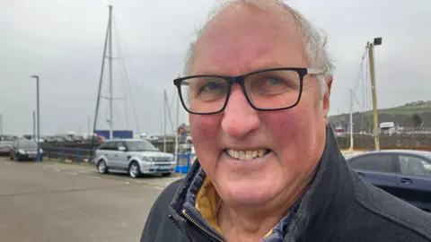 Steven Rogers, who has short white hair and wears glasses, in Whitehaven with boat masts in the distance behind him.