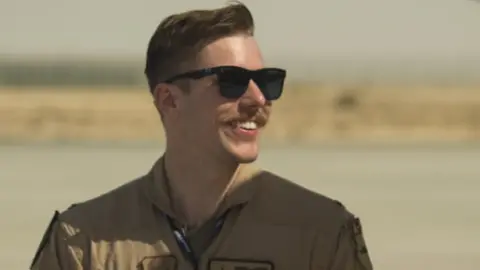 A US Air Force pilot wearing dark glasses and a moustache. He is in a brown flying suit. The man is looking away from camera and he is smiling.