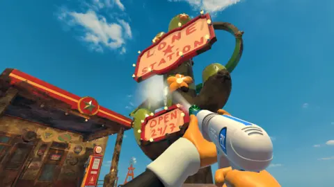 An in-game screenshot of the game PowerWash Simulator 2. It shows a first-person perspective of someone cleaning the red and white sign of a gas station outside that reads "Lone Station" and "Open 24/7" with a large green cactus as the pole. They are using a grey and blue power washer and have orange gloves. The sign is next to a gas station which is covered in dirt. 