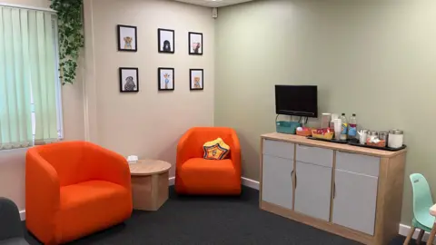 A view of a family room featuring two orange one-seater chairs with a round wooden table in between. To the right is a unit featuring three cupboards with a TV and coffee station on top.