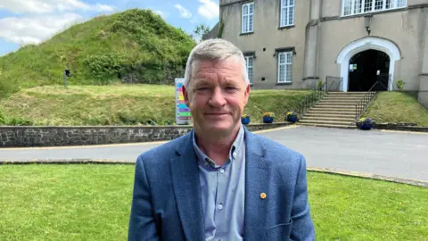 Bryan Davies looks at the camera. He has short grey hair and is wearing a blue tweed blazer with a striped blue shirt underneath. He is standing on a grass lawn with the university building behind him. In the background stone steps lead up to the black door of the stone building.