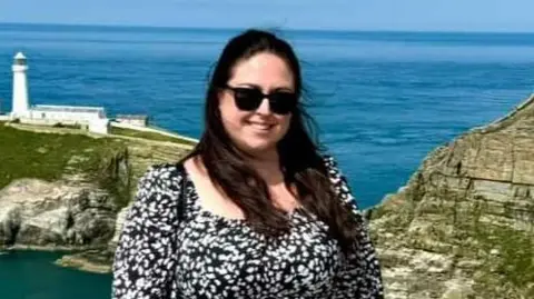A brunette woman in a black and white dress stood in front of a scenic viewpoint. In the background, we can see a white lighthouse on an island encompassed by the sea.