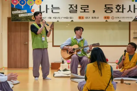 BBC/ Hosu Lee Participants sit and perform music at the temple dating retreat - one person stands holding a microphone, while another sits playing an acoustic guitar. Other attendees sit on floor cushions in a circle. A large banner with Korean text hangs behind the group.