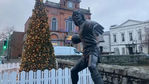 An outside shot of Newry town. A black statue of a footballer about to throw a football can be seen in front of a Christmas tree adorned with yellow lights. A big red brick building with a clock at the top and a white classical style building with pillars can be seen in the background.
