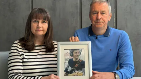 BBC Niamh Dolan, wearing a black and white striped top, and her husband Peter Dolan, wearing a blue jumper over a dark blue polo shirt, both sit looking towards the camera, holding a photo of their late son Enda, who is pictured playing guitar and smiling. 