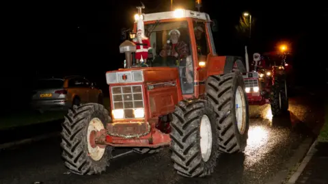 Somerset Agri Pics A red tractor in the dark with a Santa figure on the front and the driver wearing a Santa hat. Another red tractor is following close behind