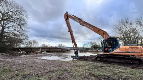 An orange crane with a large claw sits on the edge of a river bank with a low bridge in the background which is partially collapsed on the right side.