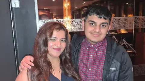 Pooja and Ronan are stood outside a restaurant. Ronan has his arm around his mother. He is wearing a red checked shirt and black jacket. She has long brown hair and is wearing a sleeveless top.
