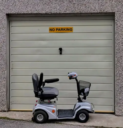 Stephen Choate A mobility scooter parked outside a garage door with a No Parking sign