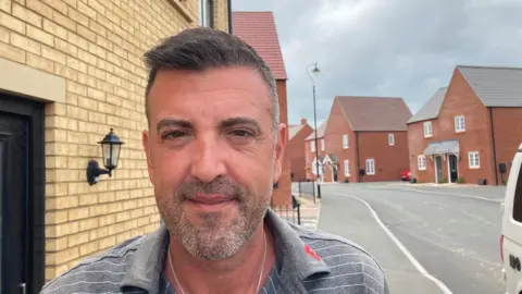 A man is standing on a street next to a house. He has short dark hair and a short beard. He is wearing a grey and white stiped T-shirt and a chain round his neck.