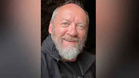 Adrian Hanna Adrian Hanna. He has a thick white beard, is bald at the top with white hair on the sides. He has blue eyes, is smiling, and wearing a black jacket with black t-shirt.