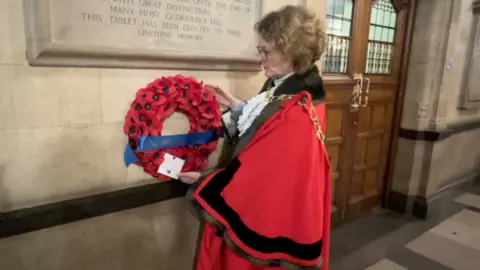 The Lord Mayor of Oxford Louise Upton, in red ceremonial robes, placing a poppy wreath at a war memorial plaque in Oxford Town Hall
