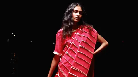 A woman wearing a pink and white striped sari stands against a black backdrop. She has straight black hair.