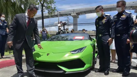 Officials in Thailand stand next to a green Lamborghini Huracán Spyder. Reporters with cameras can be seen in the background.