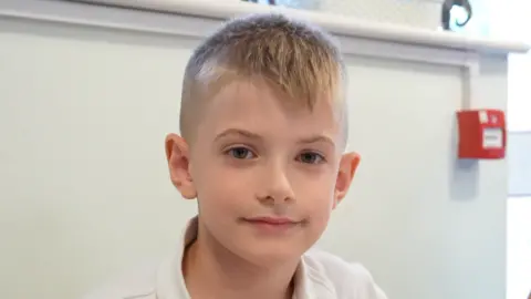 Qays Najm/BBC A young boy with blonde hair that rests over his forehead. He is wearing a white school polo top. 