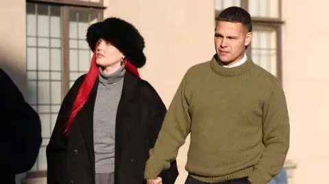 Tyron Frampton, a man with short brown hair and wearing a green knitted jumper, arriving at Oxford Crown Court on a sunny day with his wife Anne-Marie, a woman with pink hair and wearing a black hat and coat.