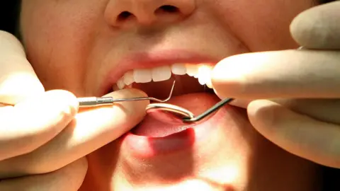 An open mouth. There are hands with gloves on over the mouth holding dentist equipment.