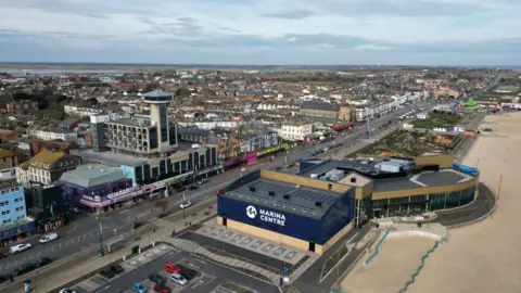 A drone shot of Marine Parade in Great Yarmouth looking north along the beach, with a tower, leisure centre and seafront arcades in view on a cloudy day.