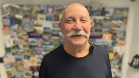 Jon Cleave is smiling at the camera. He has a bald head and big bushy moustache. He is wearing a blue T-shirt and has a fluffy microphone attached to his collar. Behind him is a panel of brightly-coloured cards.