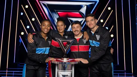 BBC/Hungry Bear Two women and two men with their hands on a trophy. They are wearing tracksuits and are standing on a stage with lights in the background.