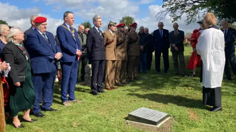 The memorial service held by Reverend Pat Gifford. It is a sunny day and people from the army, locals and local leaders are in attendance.