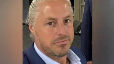 Other Ryan is taking a selfie and smiling at the camera. He is wearing a blue shirt and and suit jacket and his blonde/grey hair is scraped back.