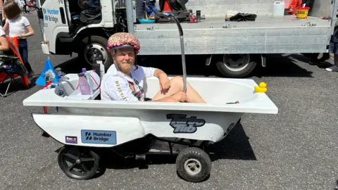 BBC A man wearing a white T-shirt, shorts and a colourful helmet sat inside a kart made out of a bath. 