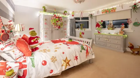 Kirsty Anderson Bedroom decorated with a Grinch Christmas theme, featuring festive bedding, green and red ornaments, and fairy lights. A Grinch toy sits on top of the bed wearing a Santa suit. 