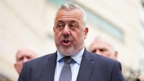 pa Neil McGuinness has grey hair and wears a suit and tie.