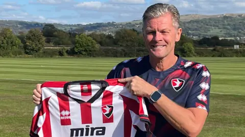 Steve Cotterill stands on a football pitch as he smiles and holds up the red and white Cheltenham Town jersey. He has grey hair and is wearing navy and red kit. Rolling hills can be seen behind him. It is a sunny day.