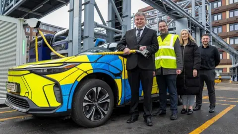Essex Police Four people stand in front of an Essex Police electric vehicle. From the left they are Jason Tyrrell, assistant head of fleet for Essex Police (who wears a black suit with grey shirt and dark blue tie), Andy Gwilliam, Head of Product at First Bus (who wears a suit with a yellow high-visibility jacket on top), Sergeant Harriet Clarke from Essex Police (who wears light grey trousers and a long black puffa jacket over the top) and Martin Fitzjohn, Strategic Commercial Solutions Manager for Seven Forces who wears black trousers and a black jacket.
